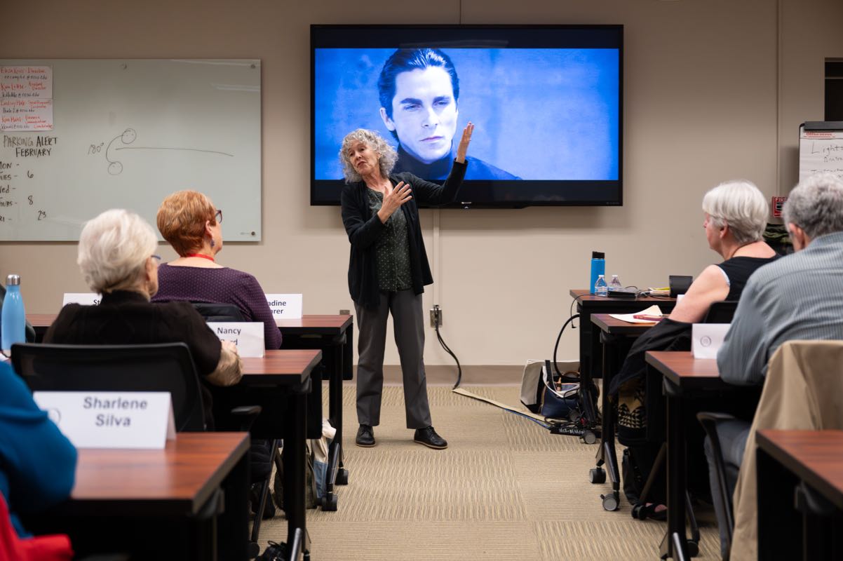 An instructor speaks in front of a large TV while lifelong learners sit at desks and listen to the presentation