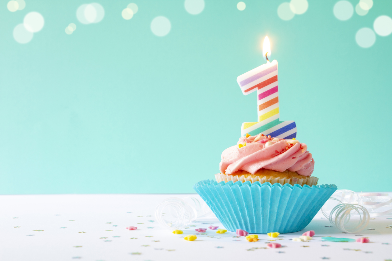 Vanilla buttercream cupcake with pink icing, sprinkles and 1st birthday candle, on a light blue background.